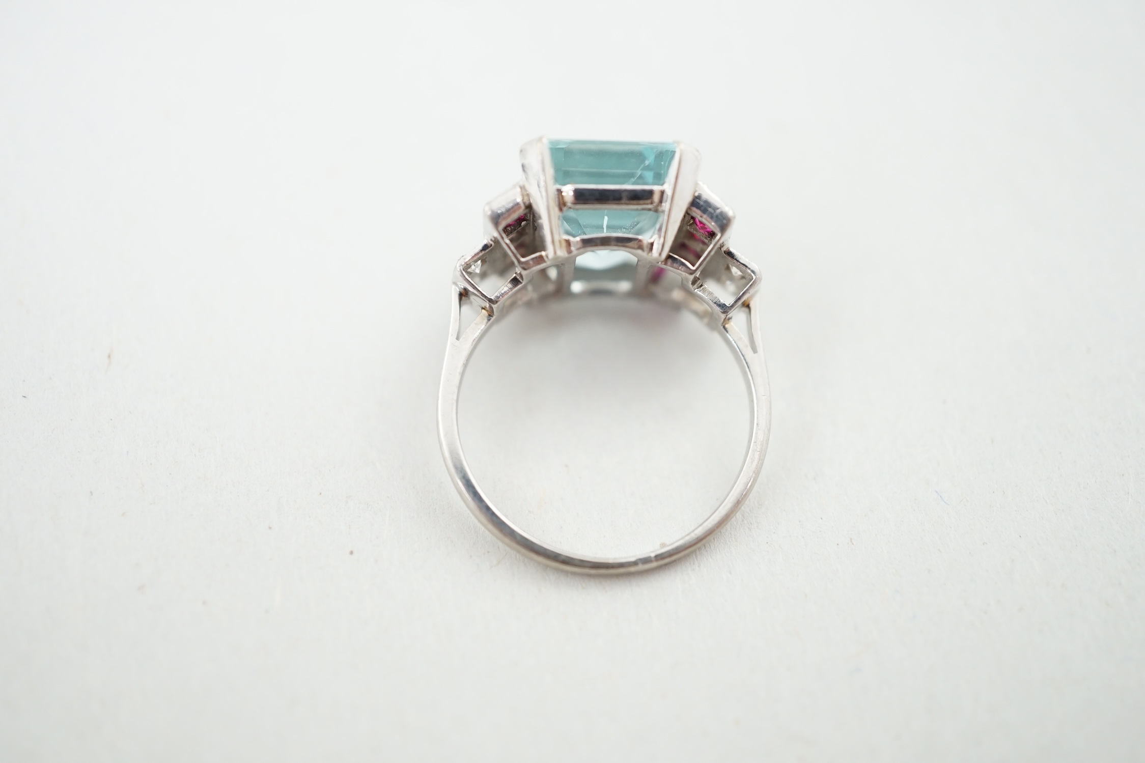 An Art Deco style platinum? and single stone emerald cut aquamarine set dress ring, with millegrain set ruby and diamond cluster stepped shoulders